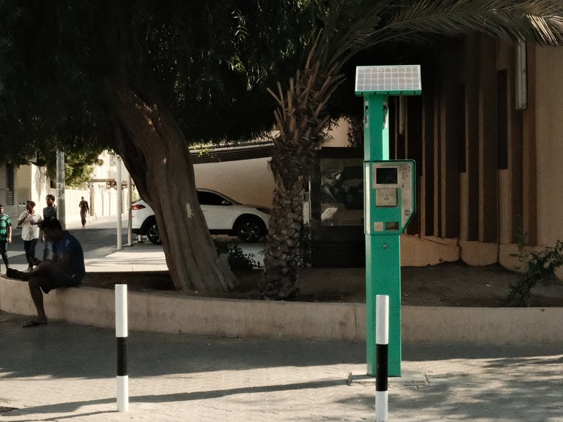 A green parking meter beside a palm tree in an urban setting with a car and a person in the background.