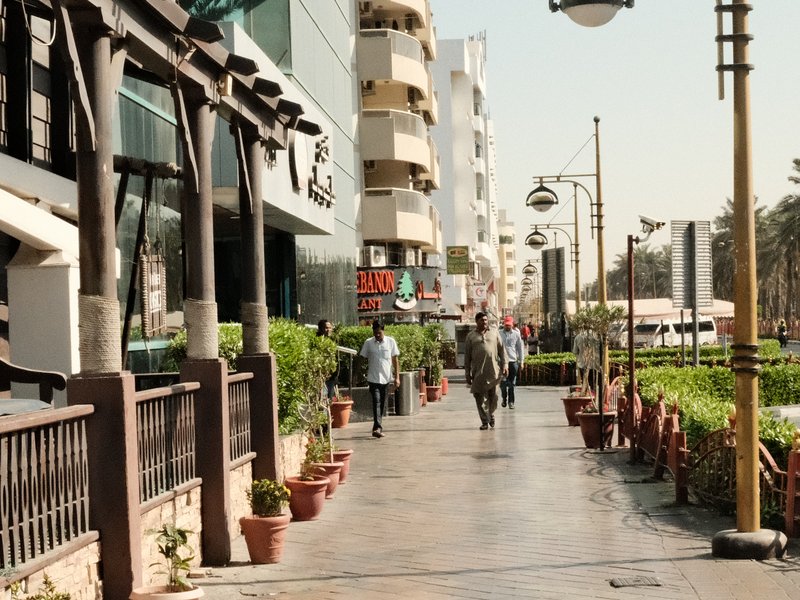 A bustling urban walkway lined with shops and greenery, featuring pedestrians strolling under bright sunlight.
