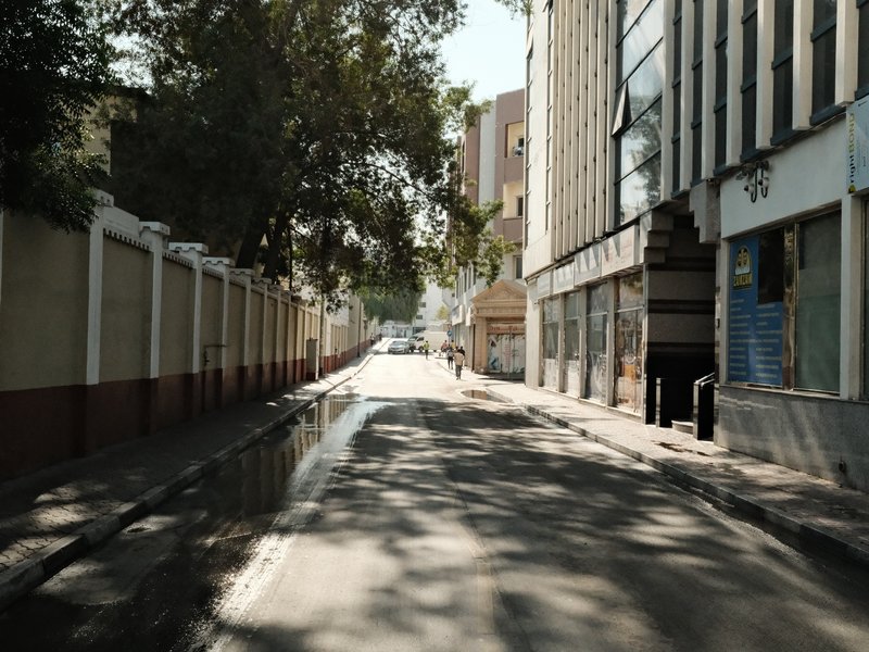 A quiet urban street with buildings on either side and a puddle reflecting the sunlight.