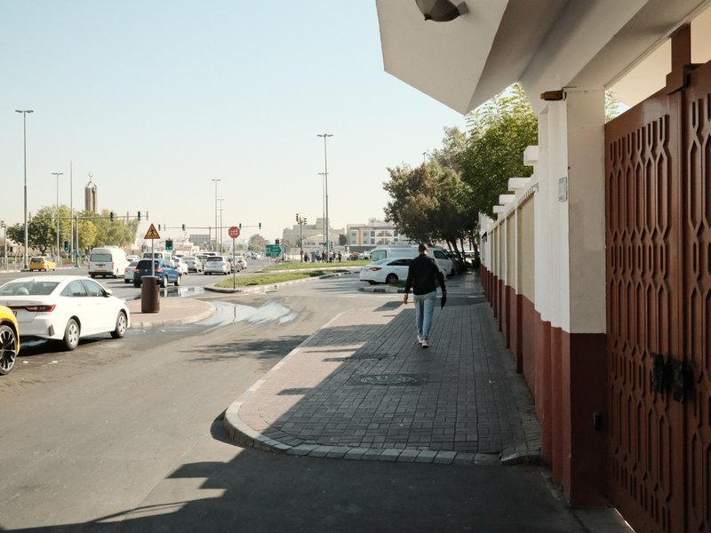 A person walking on a sidewalk beside a road with parked cars and a wall in a sunny urban setting.