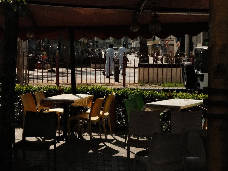 Outdoor café scene with empty tables and two figures walking in the background.