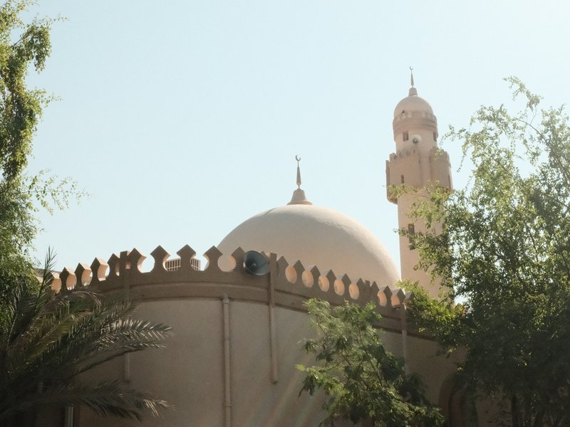 A mosque with a dome and minaret partially obscured by greenery under a clear sky.