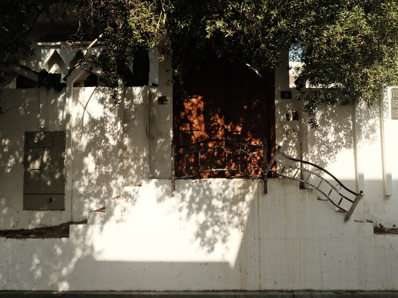A partially collapsed white wall with a wooden door and shadows from nearby trees.