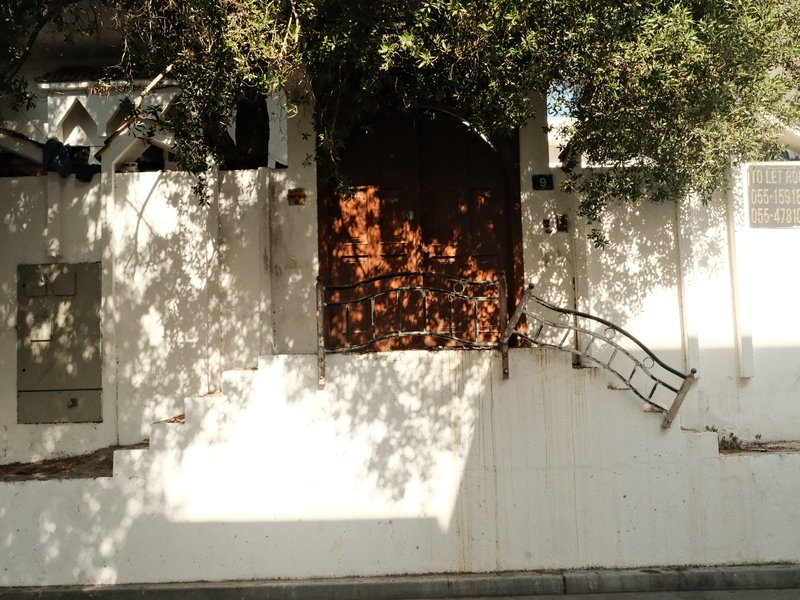 A white wall with a wooden gate partially open, surrounded by greenery and shadows.