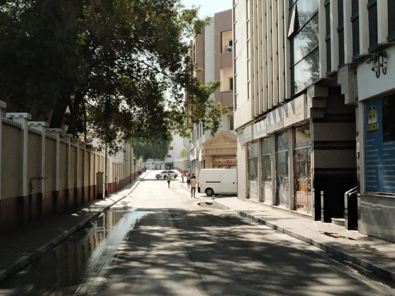 A quiet urban street with buildings on either side and a few vehicles in the distance under bright sunlight.