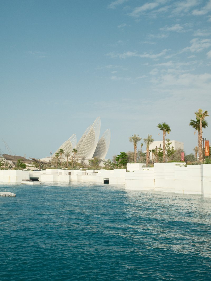 View of a modern architectural structure with palm trees beside a calm blue water body under a clear sky.