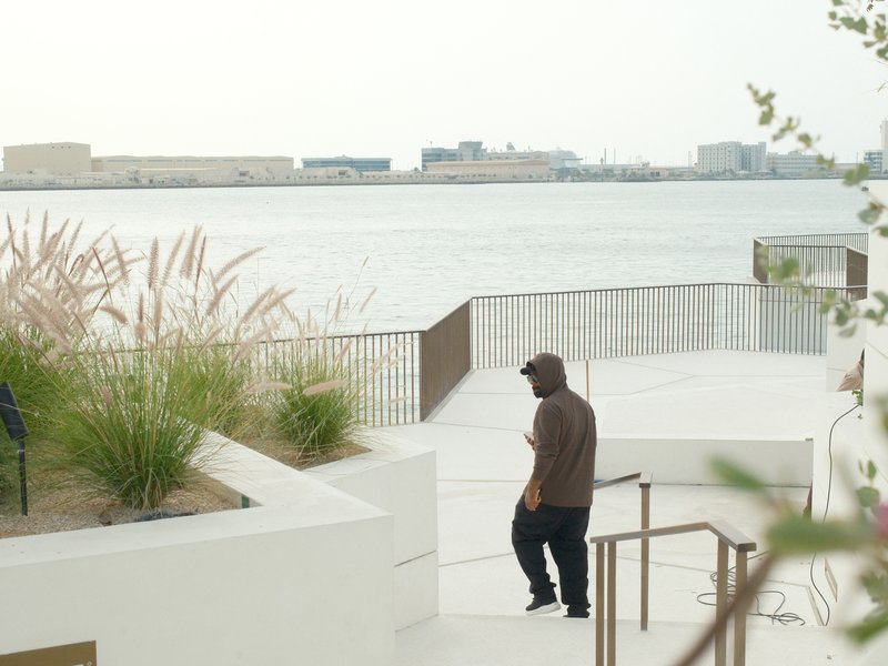 A person in a brown hoodie walks down steps near a waterfront with grass and buildings in the background.