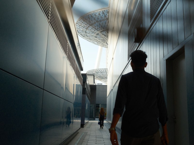 A person walking through a narrow urban alley with modern architecture and a dome structure visible above.