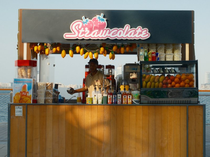 A colorful fruit stand with a vendor, displaying various fruits and drinks against a coastal backdrop.