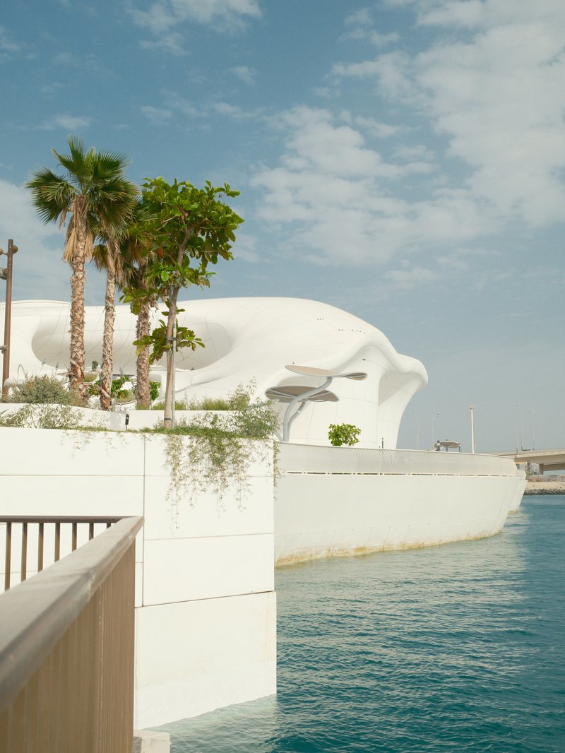 A modern architectural structure beside a calm body of water, with palm trees and a clear sky.