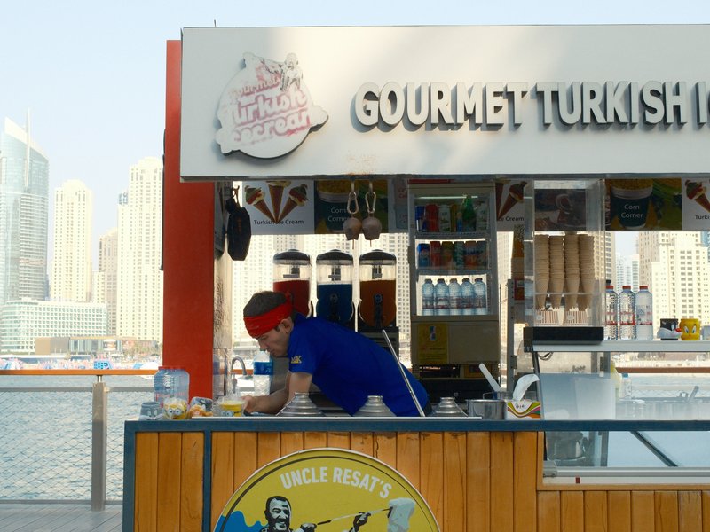 A vendor preparing food at a gourmet Turkish food stall by the waterfront with city skyline in the background.