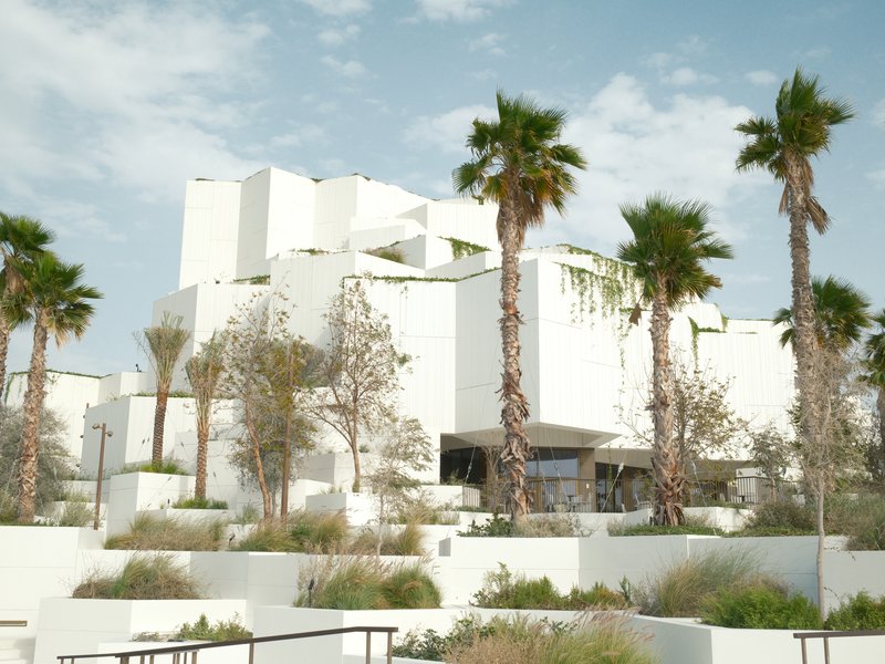 Modern white architecture surrounded by palm trees and greenery under a blue sky with clouds.