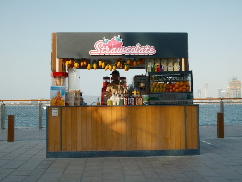 A colorful food stall with various fruits and drinks, set against a blue sky and water backdrop.