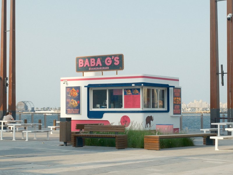 A colorful food stall named Baba G's by the waterfront with a clear sky and distant city skyline.