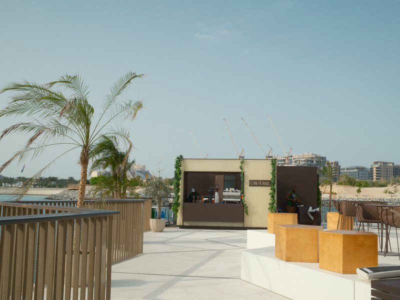 A beachside café with palm trees and wooden seating, overlooking the water on a sunny day.