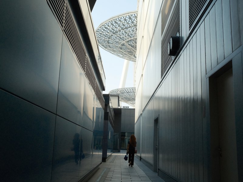 A narrow alleyway between modern buildings leading to a futuristic structure above.