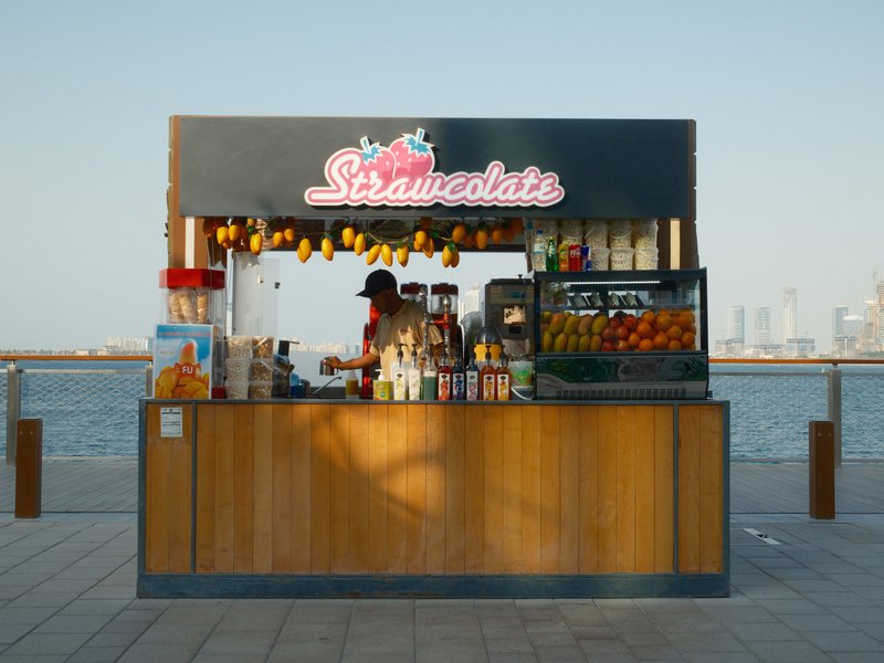 A colorful fruit stand by the water, with a vendor serving drinks and fruits under a clear sky.