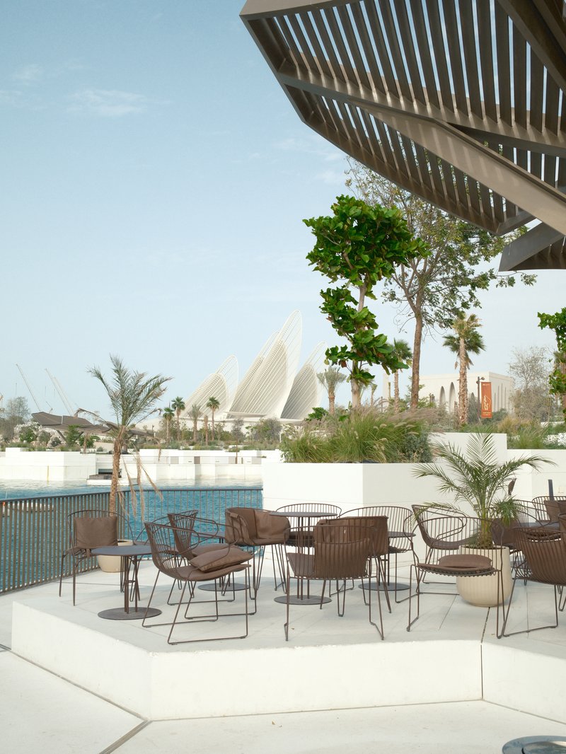 Outdoor seating area by a pool with modern architecture in the background and greenery around.