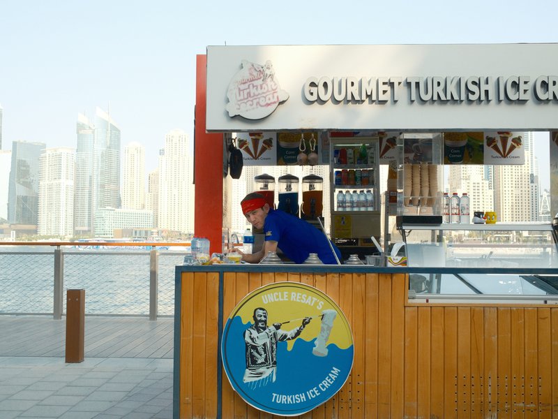 A vendor at a Turkish ice cream stand with a city skyline in the background.