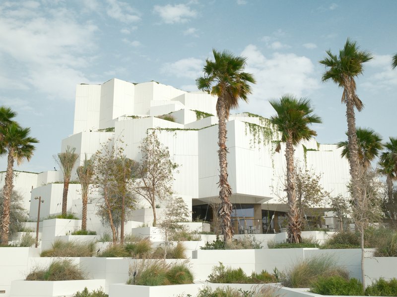 Modern architecture with white geometric structures surrounded by palm trees and greenery under a clear sky.