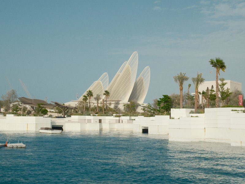 Modern architectural structure resembling sails near a body of water with palm trees and clear sky.
