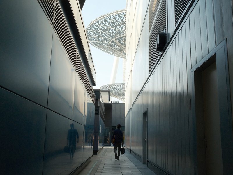 A person walking through a narrow alley between modern buildings, with a futuristic structure visible above.