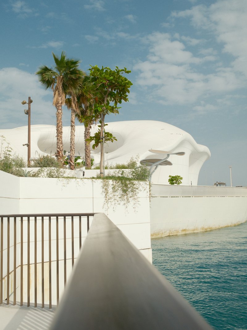 A modern architectural structure with palm trees and a railing overlooking a body of water under a blue sky.