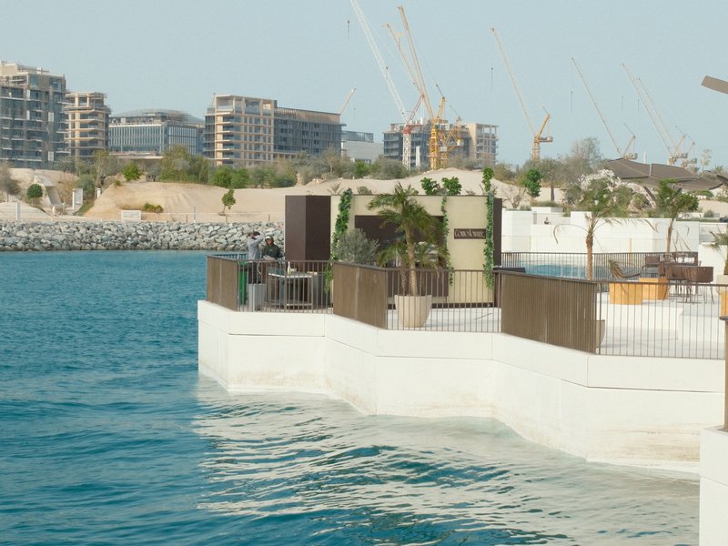 A modern waterfront structure with greenery, overlooking a calm blue sea and distant city skyline.