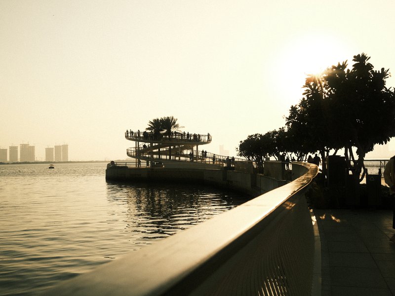 A serene waterfront scene with a winding path, trees, and a distant city skyline at sunset.