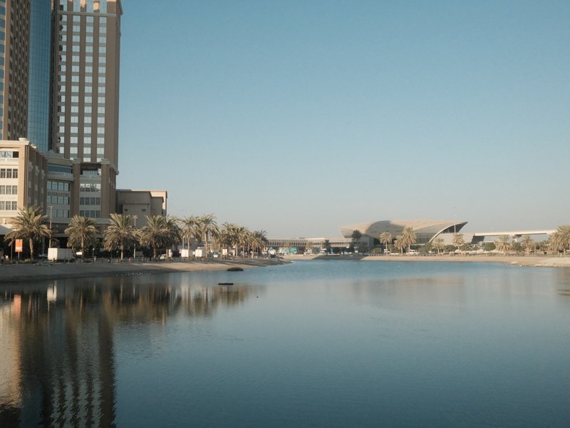 A calm river reflecting buildings and palm trees under a clear blue sky.
