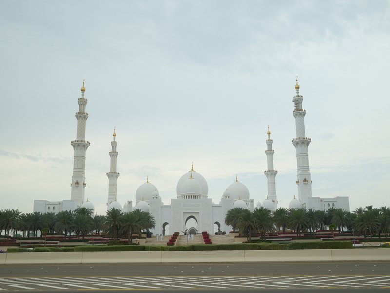 A grand white mosque with multiple minarets and domes, surrounded by greenery under a cloudy sky.