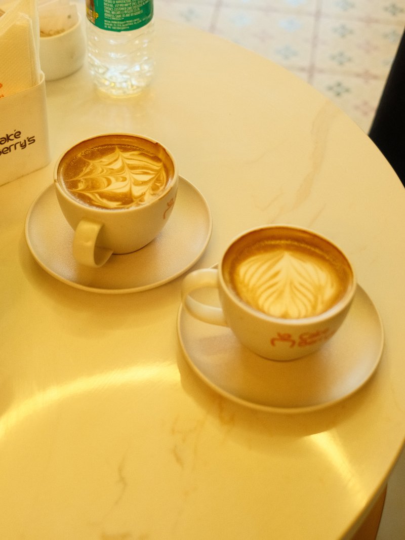 Two cups of latte art on a marble table with a water bottle in the background.