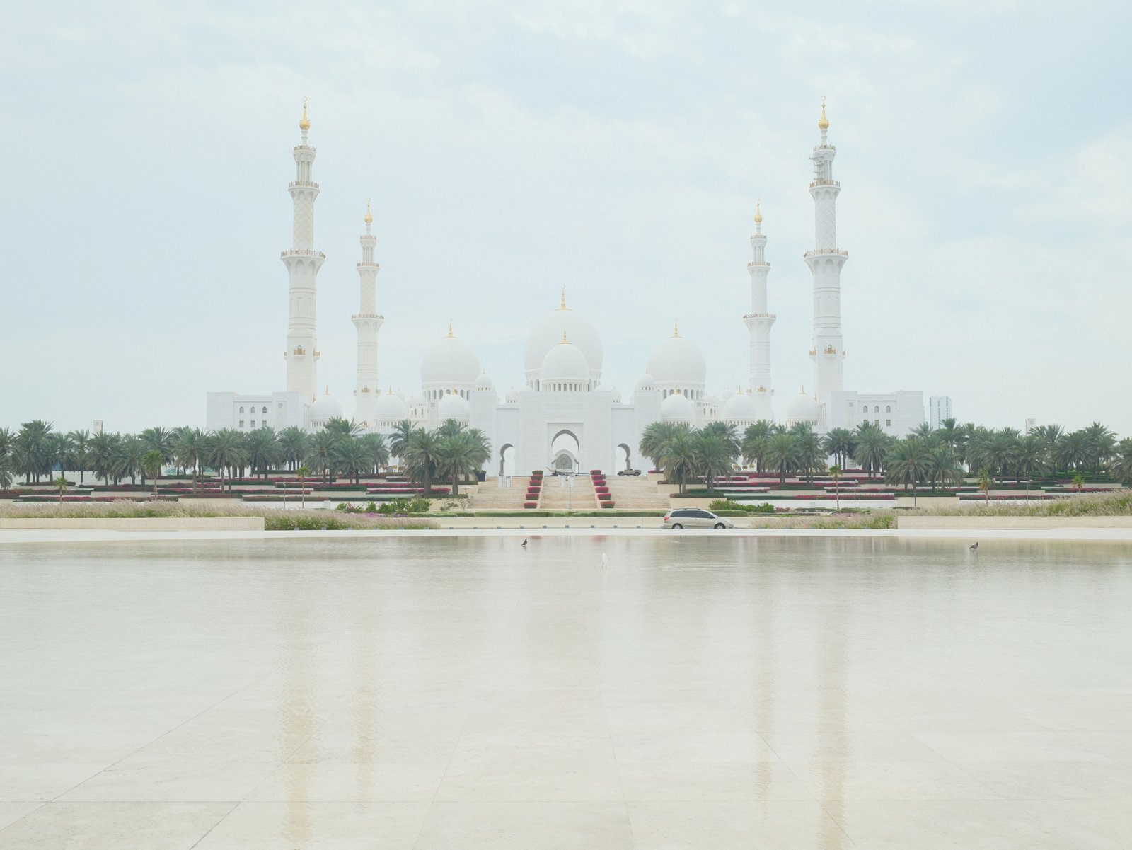 A grand white mosque with multiple minarets reflected in a calm water surface under a cloudy sky.