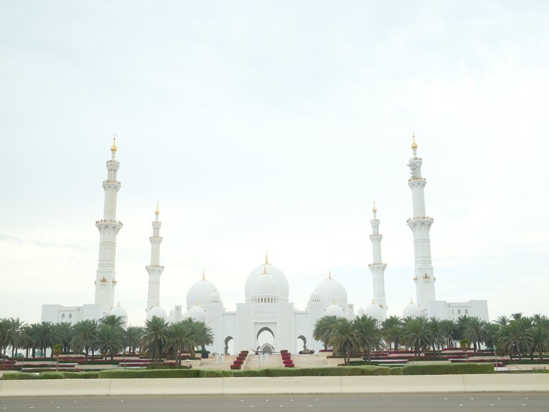 A grand white mosque with multiple minarets and domes, surrounded by palm trees and a cloudy sky.