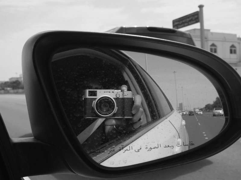 A black and white photo capturing a camera reflected in a car's side mirror with a road in the background.