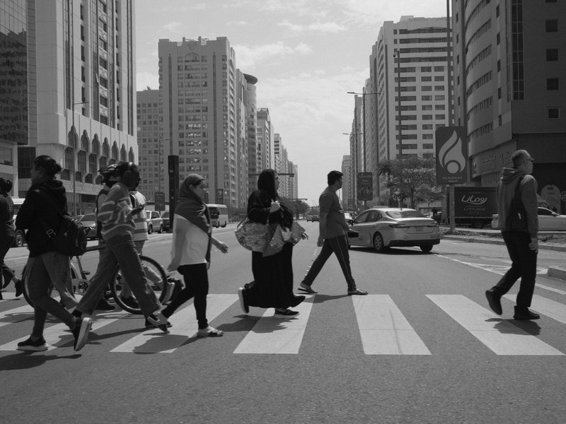 A group of people crossing a busy urban street with tall buildings in the background under a clear sky.