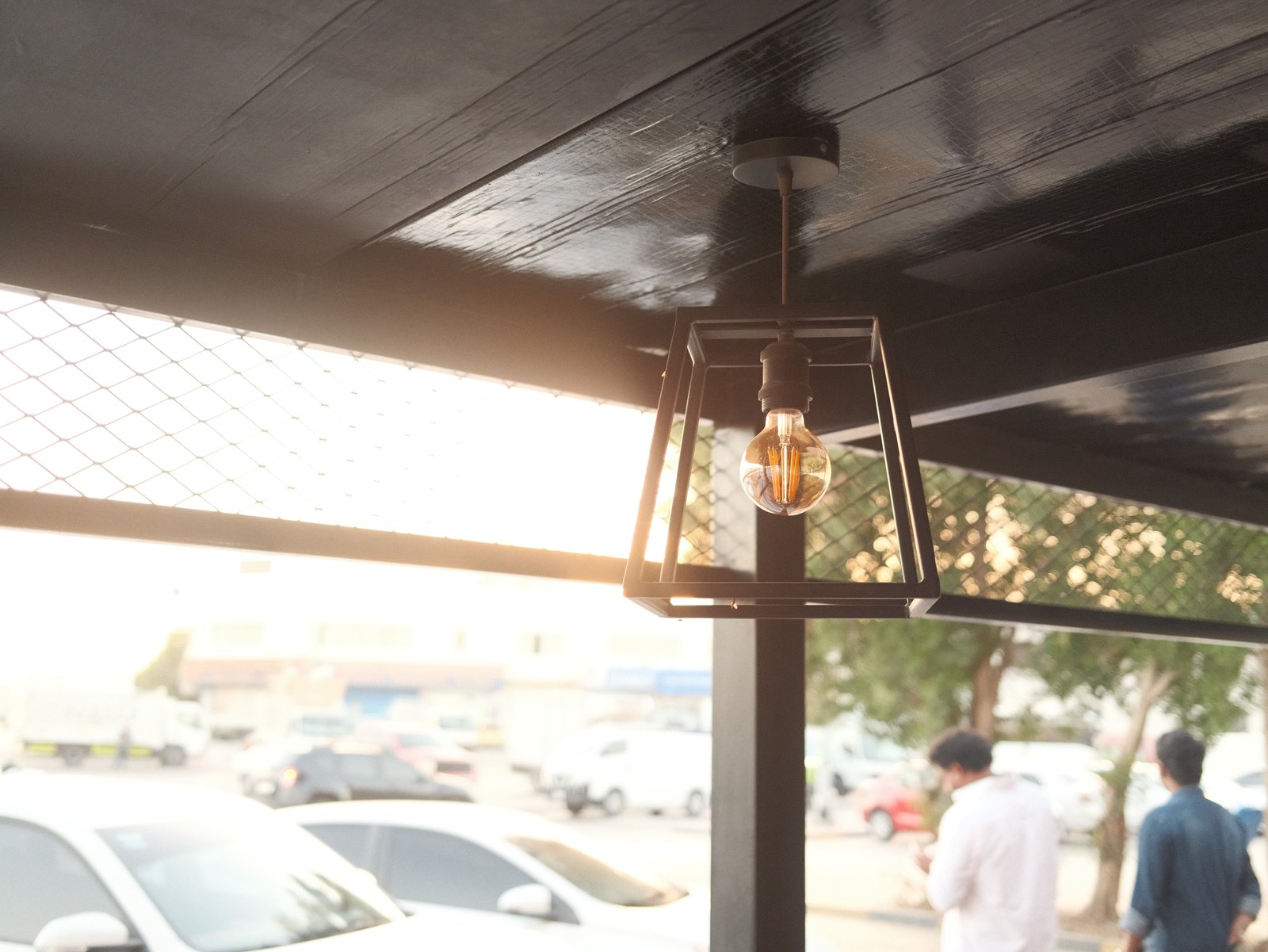 A hanging light fixture with a warm glow, set against a blurred outdoor scene with people and cars.