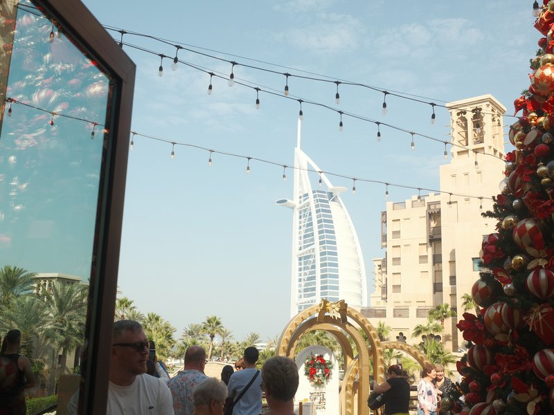 A bustling outdoor scene with holiday decorations and the Burj Al Arab in the background under a clear sky.
