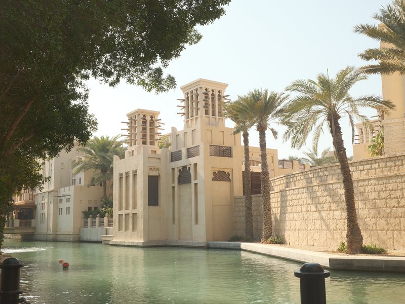 A serene view of a traditional Middle Eastern building beside a calm waterway, surrounded by palm trees.