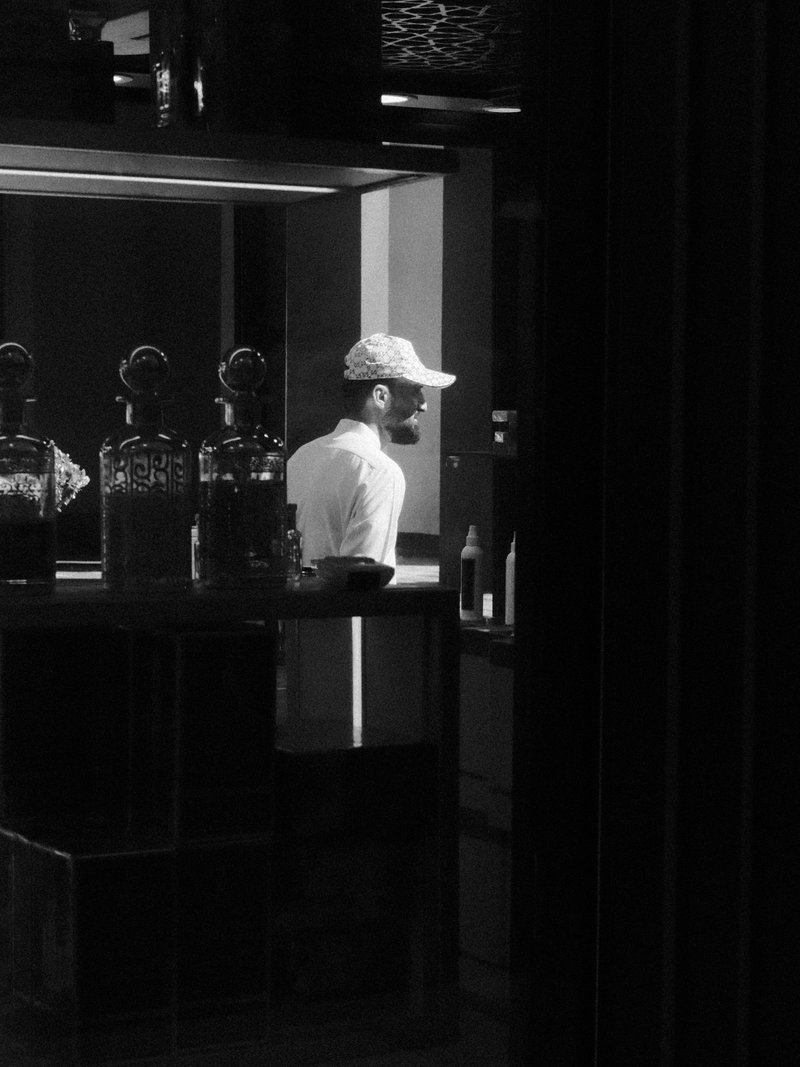 A man in a cap stands behind a bar, illuminated by soft light, creating a contemplative atmosphere.