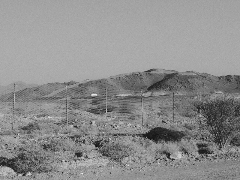 A barren desert landscape with distant mountains and a fence line under a clear sky.