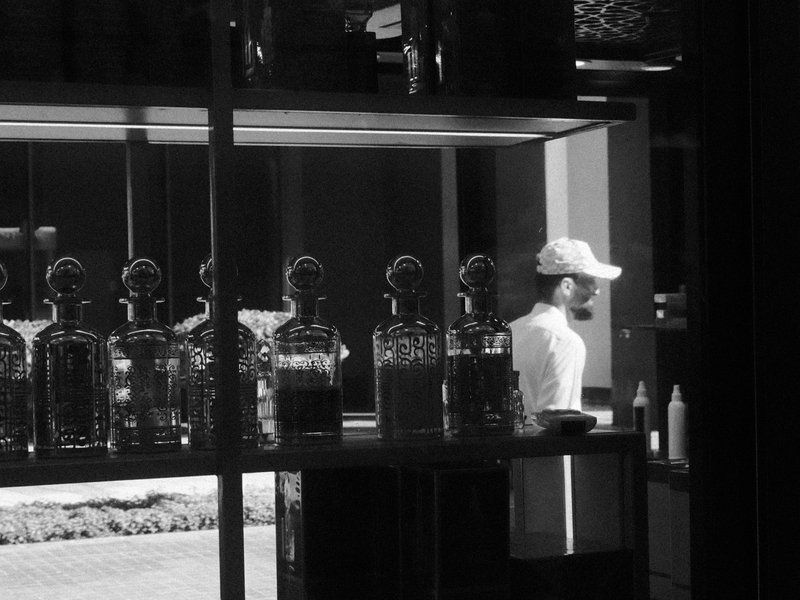 A person in a cap stands behind a glass display filled with bottles, captured in black and white.