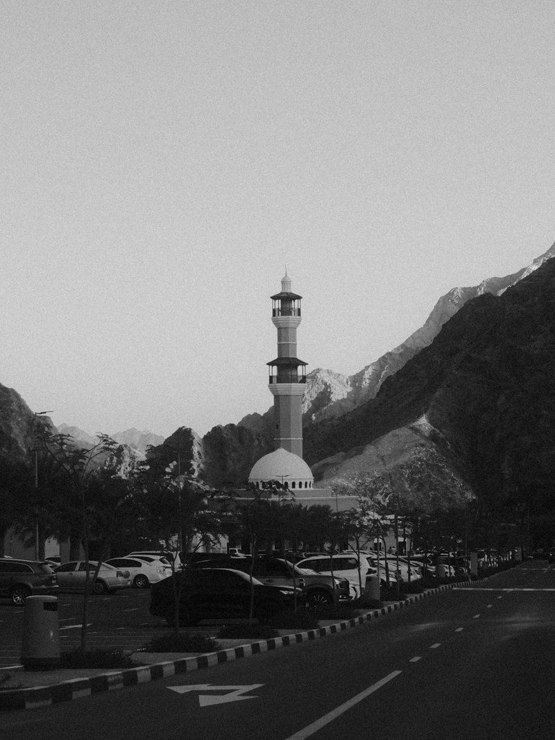 A black and white photo of a mosque tower surrounded by mountains and parked cars on a street.