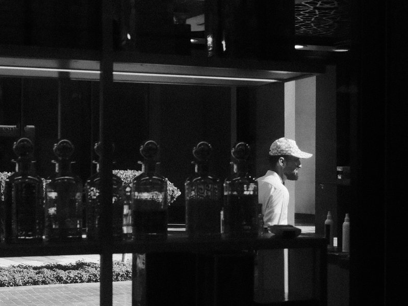 A man in a white cap stands near glass bottles in a shop, illuminated by natural light.