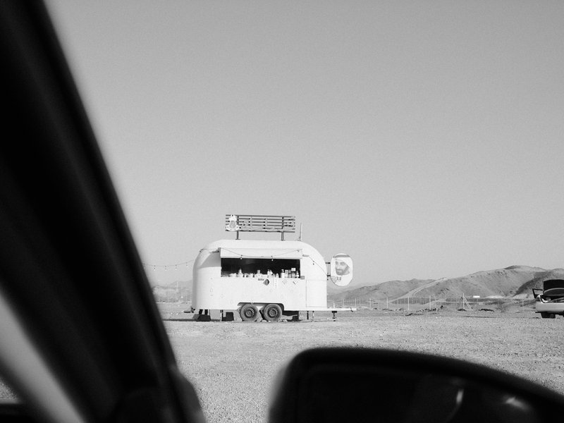 A vintage trailer in a barren landscape, viewed from inside a car with mountains in the background.