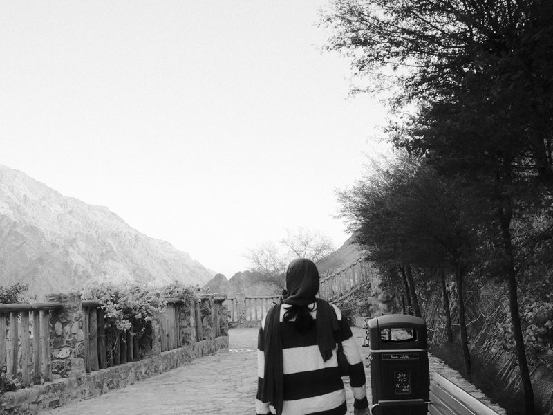 A person walking away on a path surrounded by trees and mountains in black and white.