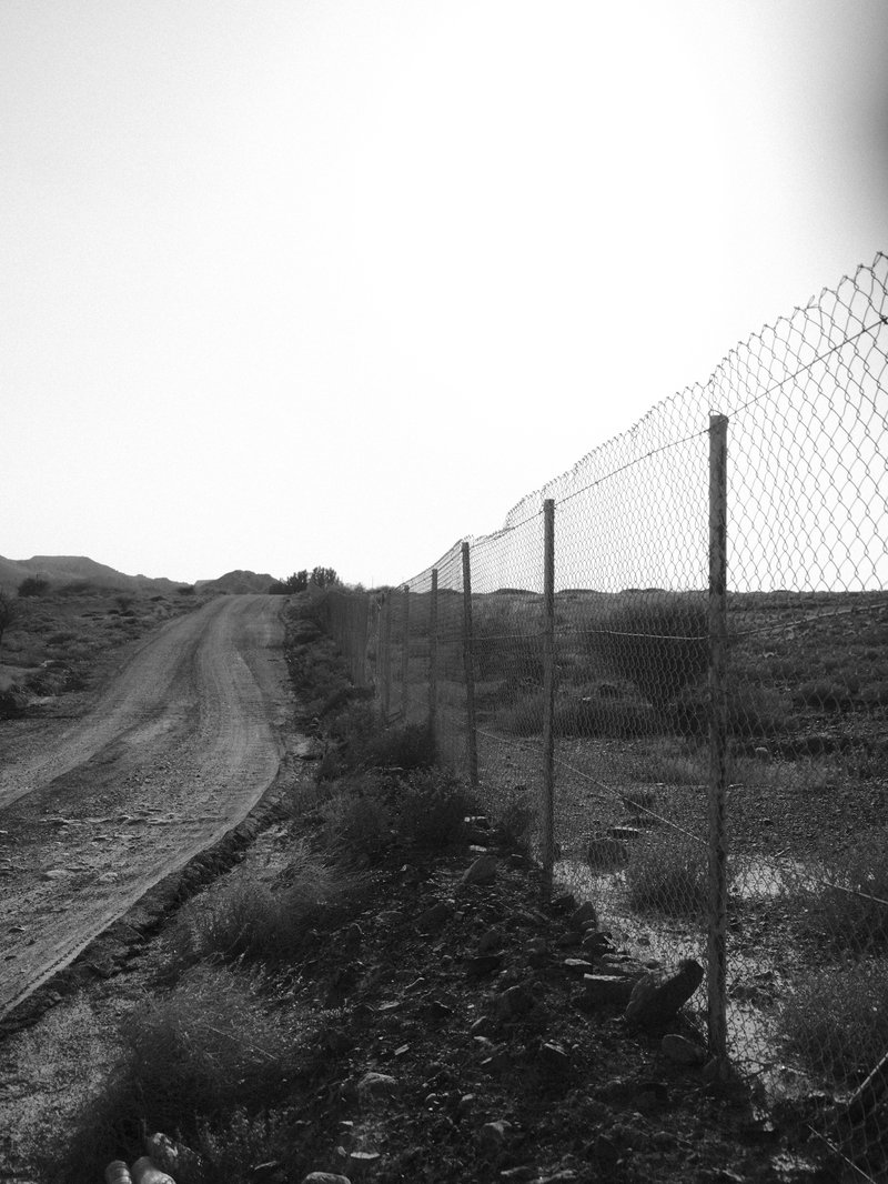 A chain-link fence along a dirt road leading into a barren landscape under a bright sky.