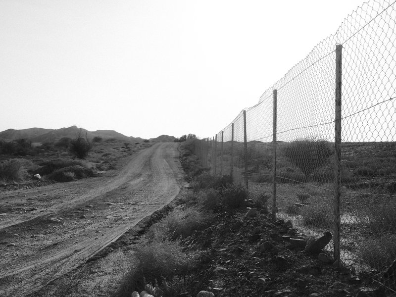 A dirt road alongside a chain-link fence in a barren landscape under bright sunlight.