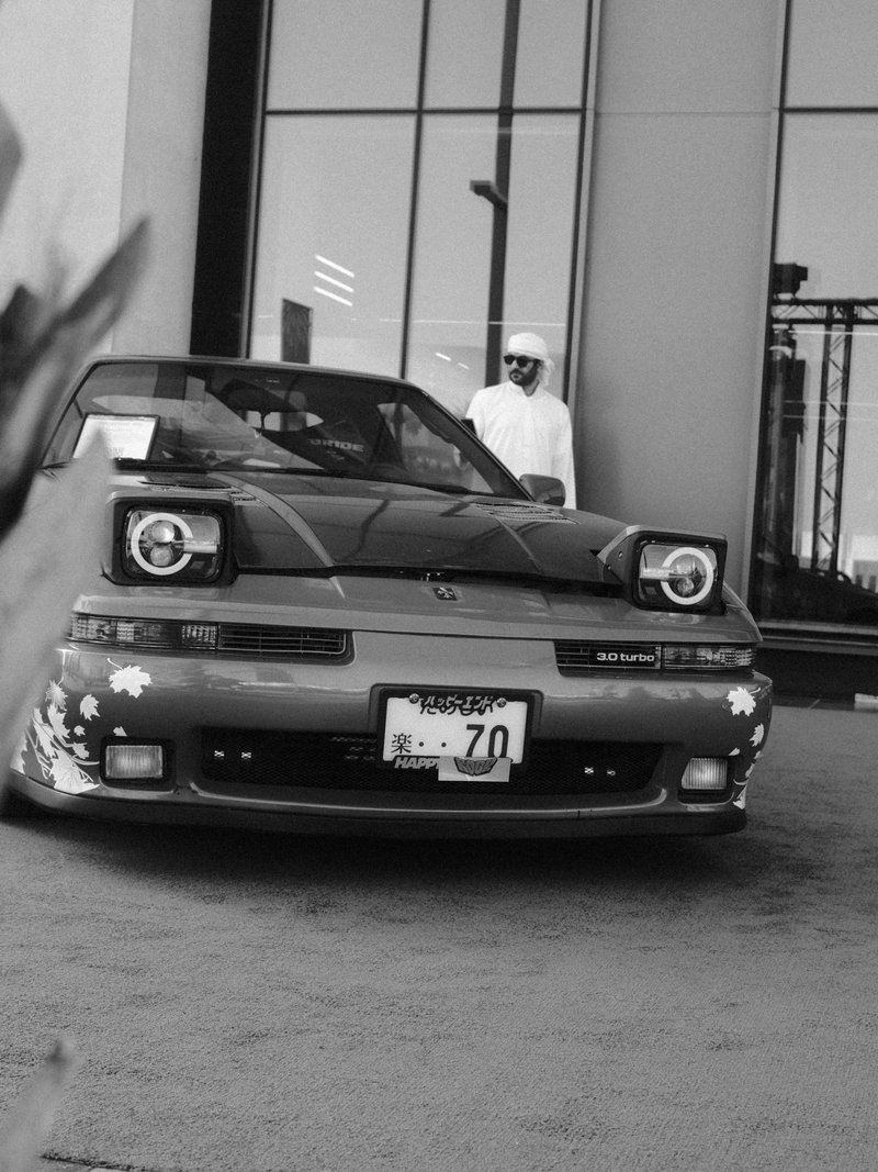A modified car parked in front of a modern building with a person in white attire standing nearby.