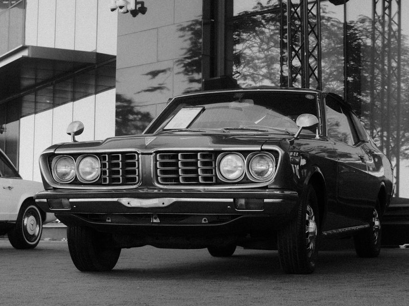 Classic black and white photo of a vintage car parked in front of a modern building.